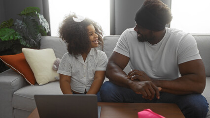 Father and daughter sitting in a living room sharing a joyful moment together with a laptop on the table, creating a warm family atmosphere indoors.