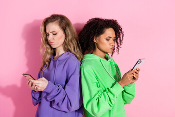 Two young women in colorful hoodies looking at smartphones against a bright pink background,...