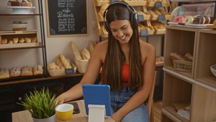 Young, attractive, brunette woman in a bakery, wearing headphones and smiling while using a tablet, seated among freshly baked bread and pastries on display
