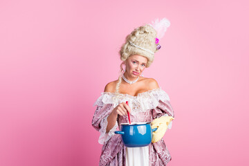 Fototapeta premium Woman in a historical costume stirring a pot, holding a creative and amusing pose with a pink backdrop