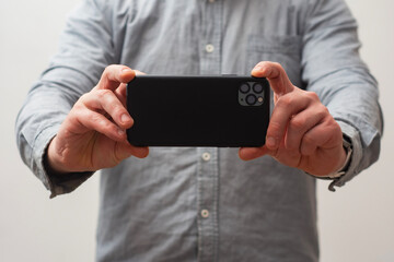 Close-up of a man wearing a gray shirt, holding a smartphone with both hands as if taking a photo or recording a video.