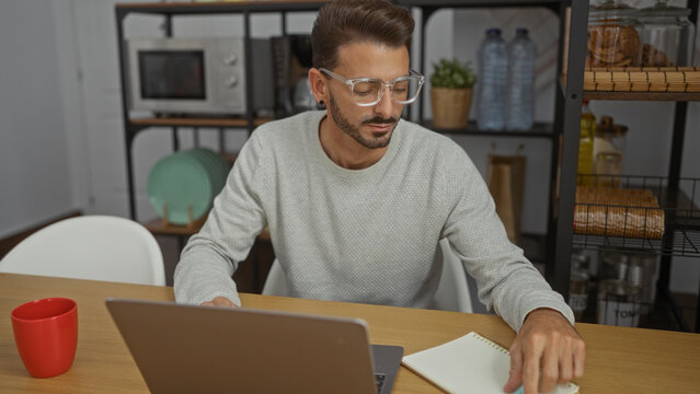 Young man working at desk in modern office interior with laptop, wearing glasses, casual sweater, focused and handsome, red mug nearby on wooden table, shelves in background