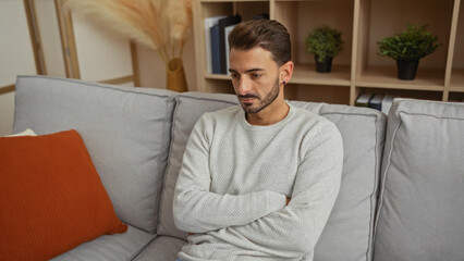 Fototapeta premium Young man with crossed arms sitting on a sofa in a cozy living room interior, showcasing a serious expression with stylish decor in the background.