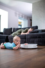 Obraz premium Cute young woman is sleeping on sofa in living room while little child is playing with robot vacuum cleaner on floor at home.