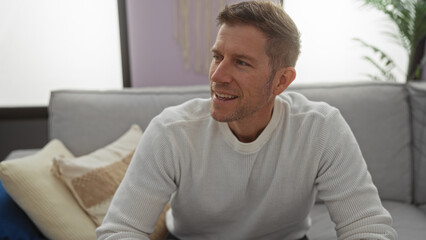 Obraz premium Young man sitting on a comfortable sofa in a modern living room, smiling confidently, creating a warm and inviting atmosphere with soft lighting around him.