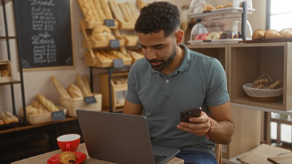 Fototapeta premium Young man working in a bakery, using laptop and phone, surrounded by bread and pastries with a coffee, highlighting hispanic culture in an indoor cafe setting.