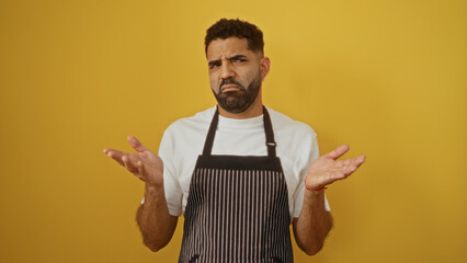 Young man in apron shrugging against a yellow background, expressing confusion or uncertainty, suitable for themes involving cooking, questions, decisions, or uncertainty in diverse contexts