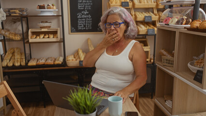 Woman with grey hair yawning in a bakery, surrounded by fresh bread and pastries, with a laptop and coffee at the table.