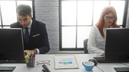 Man and woman working at computers in modern office setting with large windows, focusing intently on tasks, reflecting professional and collaborative atmosphere.