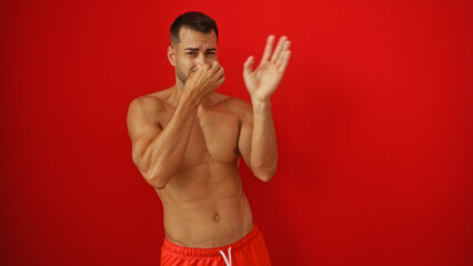 Fototapeta premium Handsome young hispanic man with a beard covering his nose due to bad smell over a red isolated background