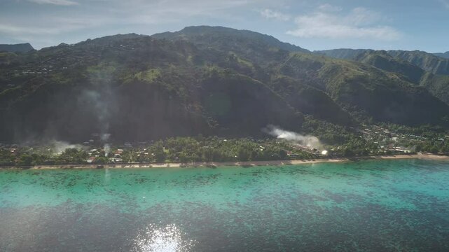 Smoke rises from a tropical village nestled on scenic coastline of ua pou, marquesas islands in French Polynesia, beauty of nature and human presence. Exotic travel vacation getaway. Drone flight