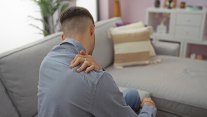 Obraz premium Young man sitting on a couch in a living room holding his back in discomfort, emphasizing a home setting with contemporary decor and a relaxed ambiance.