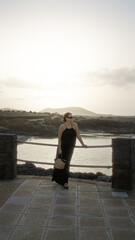 Young woman in a black dress standing by the beach in famara, lanzarote, canary islands, with mountains and the sea visible in the background at sunset.
