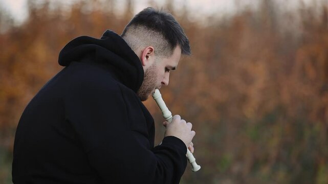 A young man in a black sweatshirt plays on panpipe enchanting melodies in the autumn park
