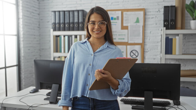Woman working in a modern office holding a clipboard, showcasing a professional setting with desks, computers, and a bulletin board, reflecting a corporate workspace.