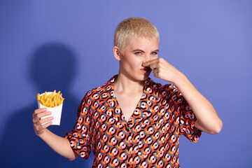 Young woman with short blond hair holding french fries and expressing dislike in front of a purple...
