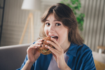 Young woman enjoying a delicious burger at home with a cheerful expression while relaxing in a modern cozy living room