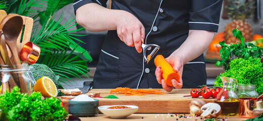 Private cook peels carrots with vegetable peeler. Cozy kitchen with wooden table, kitchenware,...