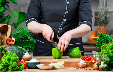 Chef blogger cuts green paprika. Cozy kitchen with wooden table, kitchenware, vegetables, herbs and ingredients for cooking. Healthy vegan eating, culinary, recipes, food blogging