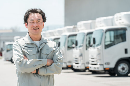 物流センターの駐車場に並ぶトラックの前にいる作業服姿の腕組みする男性（配送業・運送業・宅配業）
