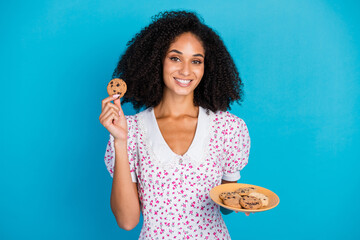 Smiling young woman holding a plate of cookies with a bright blue background, showcasing her curly hair and a summer floral dress