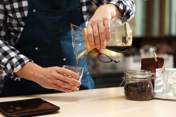 Barista pouring coffee from glass coffeemaker into cup at table in cafe, closeup