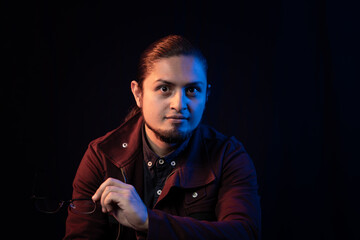 Thoughtful man with a serious expression holding glasses, illuminated by cinematic blue and warm lighting against a dark background, creating a moody portrait.