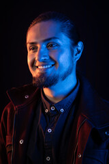 Close-up portrait of a smiling man with artistic blue and warm lighting. His confident expression and stylish jacket create a modern, cinematic look against a dark background.
