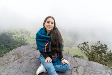 Naklejka premium Smiling young woman wearing a colorful shawl sits on a rocky cliff with a misty green valley in the background. Peaceful outdoor adventure in nature