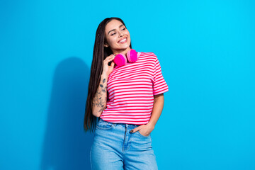 Smiling woman posing with pink headphones wearing a striped shirt and denim, standing on a vibrant blue background.