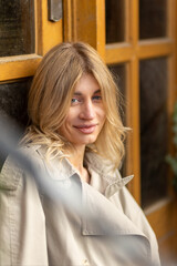 Fototapeta premium Woman with blonde hair relaxing by a yellow door while wearing a light trench coat in a cozy urban setting