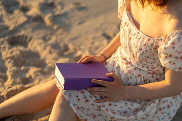Young woman enjoys reading a book on sandy beach during sunset, surrounded by warm light and serene...