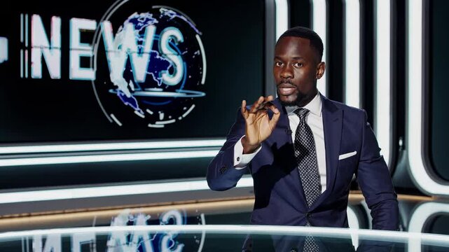 A news anchor in a suit delivers a broadcast from a modern studio. The video is shot from a front-facing angle, capturing a digital news backdrop.