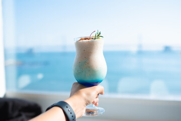 A young man is holding a glass with blue Hawaii Italian soda by the sea.