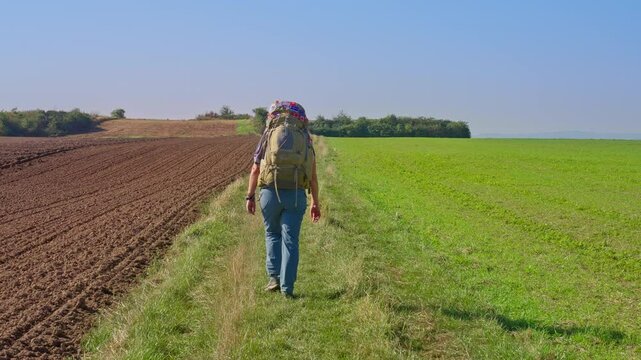 Following behind trekking hiker split between a grassy meadow and a plowed agriculture field on a sunny day, walking in slow motion