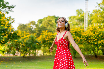 Fototapeta premium Young Woman in Retro Red Polka Dot Dress Wearing Headphones Listening and Enjoying Music in a Park Happily, Immersing with Nature