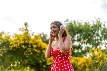 Young Woman in Retro Red Polka Dot Dress Wearing Headphones Listening and Enjoying Music in a Park...