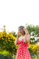 Young Woman in Retro Red Polka Dot Dress Wearing Headphones Listening and Enjoying Music in a Park...