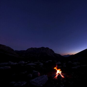 Wide shot of stars emerging over the mountain, the sky shifting from indigo to midnight blue, as a lone campfire flickers, radiating warmth.
