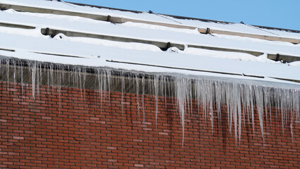 A roof covered in snow with long icicles hanging down against a brick wall under a clear blue sky.