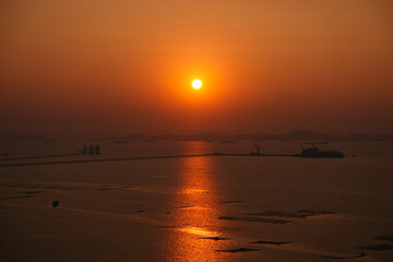Glowing orange sunset illuminating a calm sea, with an industrial harbor and distant cargo ships creating a picturesque skyline in Si Racha, Chonburi, Thailand