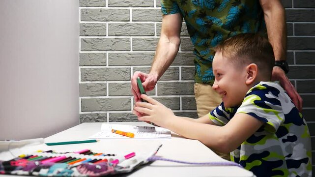 A boy plays a game on a phone sitting at the desk but his dad makes him do homework
