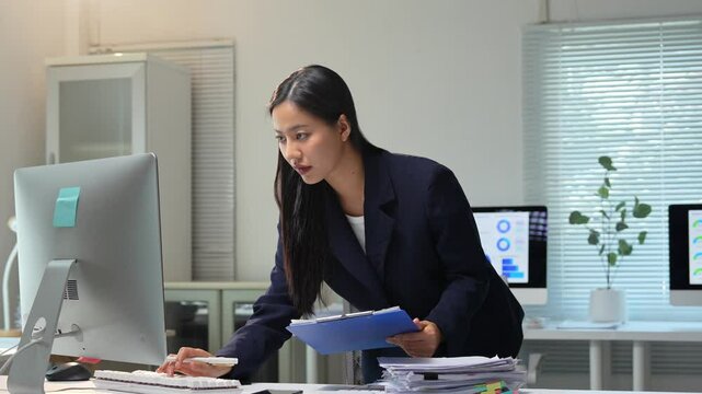 Professional businesswoman standing at desk, working on desktop computer and taking notes on clipboard in sleek, modern office environment, showcasing workplace efficiency and focused productivity