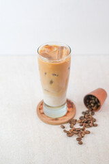 Iced Cappuccino In Tall Clear Glass With Milk And Espresso Layers Served On Wooden Coaster Beside Roasted Coffee Beans On Light Textured Surface.