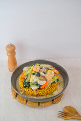 Savory Ifumie With Shrimp, Fresh Vegetables, And Flavorful Thick Gravy Presented In Grey Bowl On Wooden Trivet With Pepper Grinder Behind.