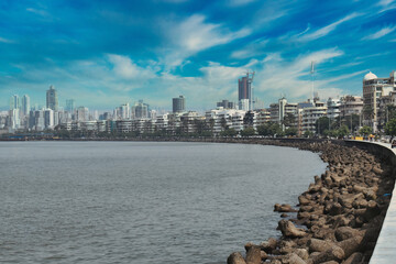 A vibrant coastal promenade features the city skyline with modern buildings and natural rocky shores under a blue sky
