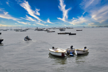 Multiple boats are anchored in a tranquil harbor as the sunset paints the sky with vibrant clouds and soft light over the water
