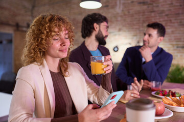 Businesswoman drinking orange juice and using smartphone during office break with colleagues chatting