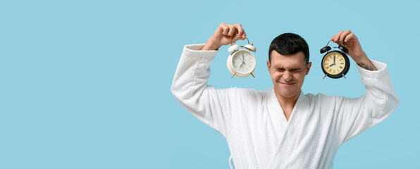 Portrait of sleepy young man in bathrobe suffering from loud noise of alarm clocks on blue...
