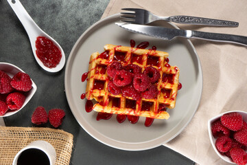 A golden waffle topped with fresh raspberries and raspberry sauce drizzling over it. The crispy...